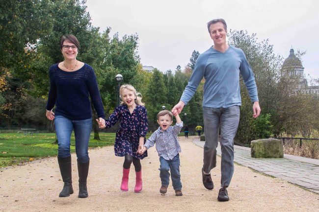 Family photo portrait at Capitol Lake in Olympia, Washington. Washington State Capitol in background. Mother, father, young daughter and son running in photograph. Family portrait session.